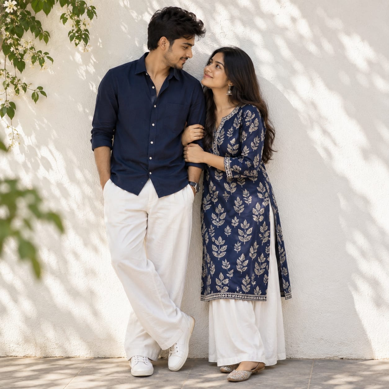 Indian couple in coordinated navy and white outfits standing against a textured wall with soft leaf-shadow patterns